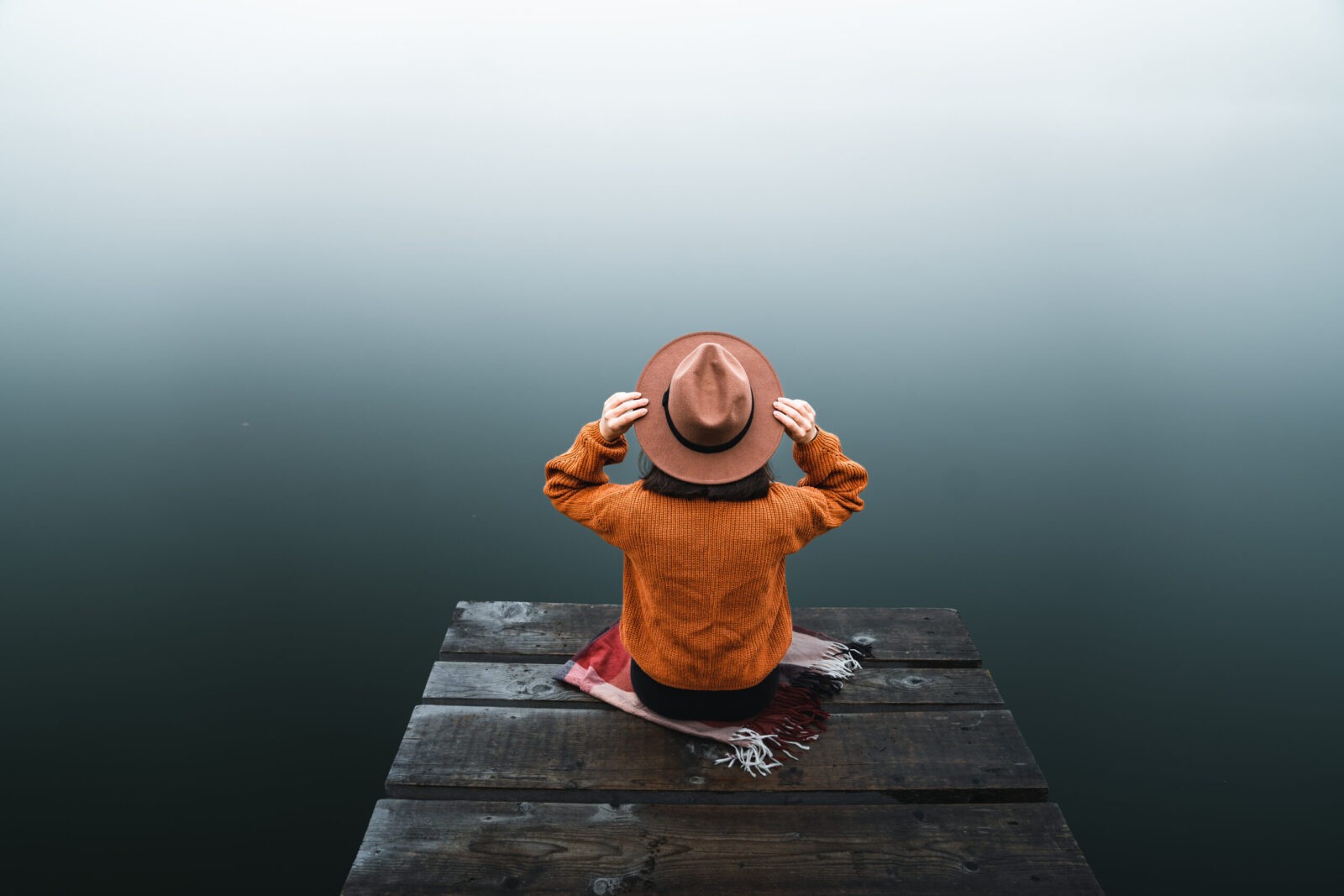 Serene scene of a woman with hat and orange sweater sitting on wooden dock facing foggy lake.