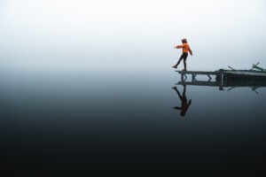 Person carefully walking on edge of wooden dock over reflective water.