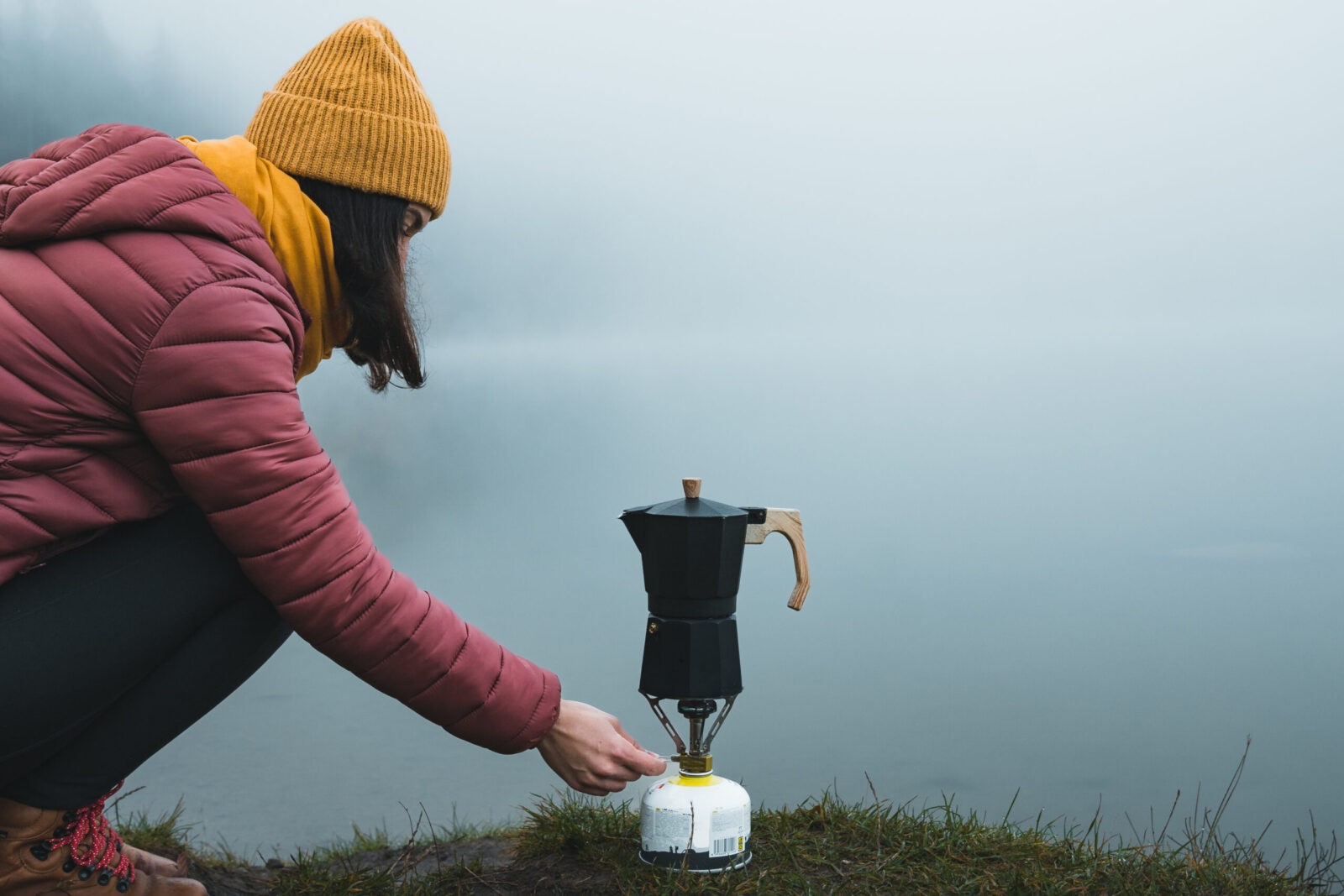 Hiker woman preparing a moka pot coffee outdoor, by a foggy lake.