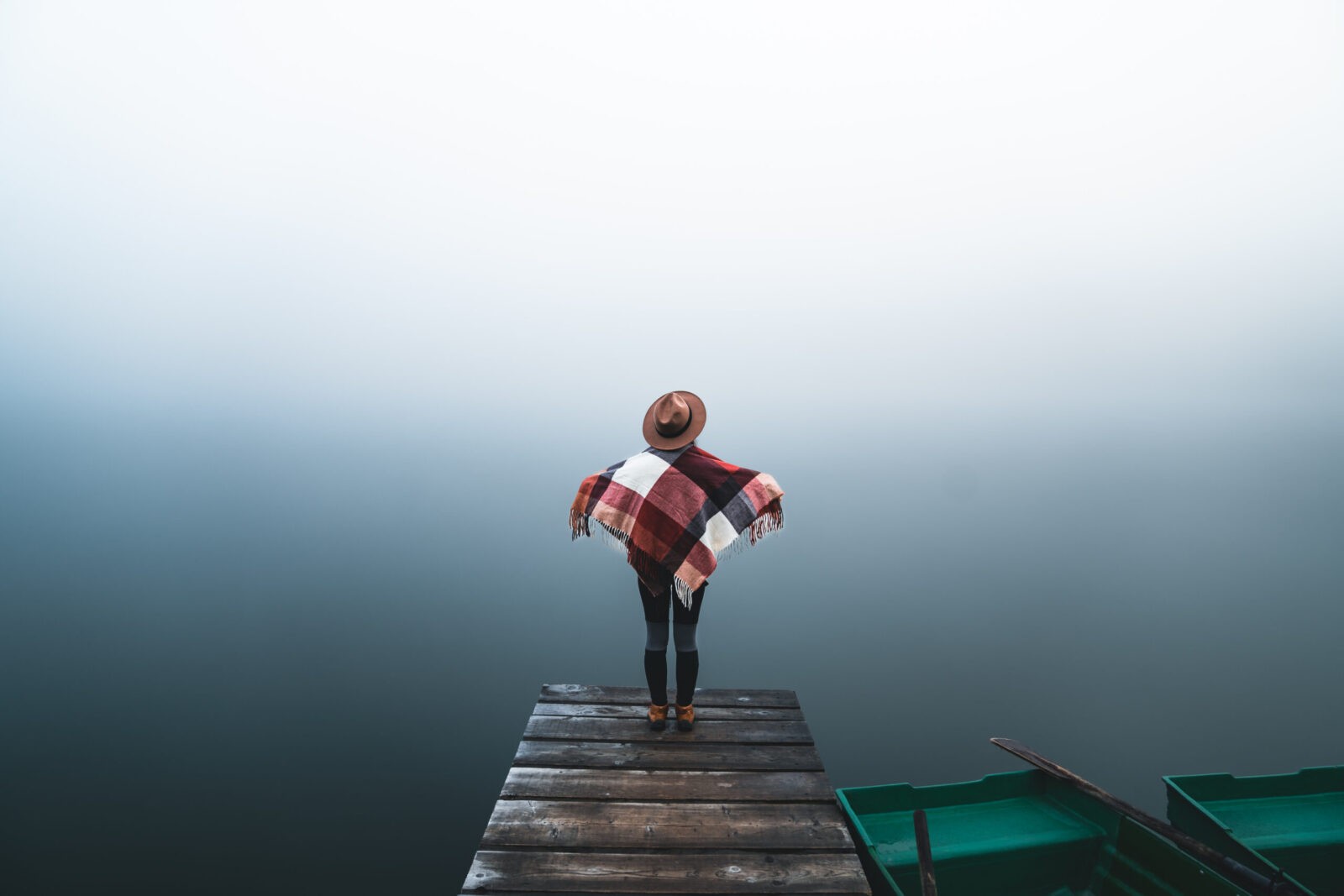 Back view of person in hat and poncho on wooden dock overlooking foggy lake.